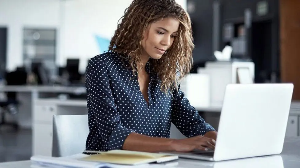 A woman in a blue polka dot shirt is working on her laptop.