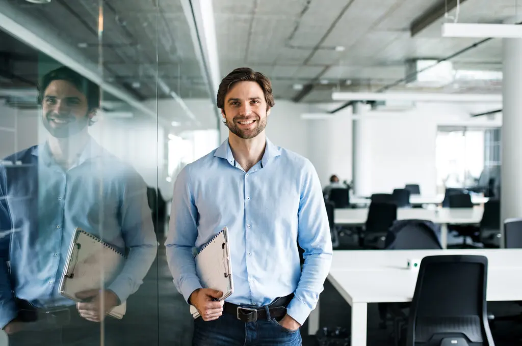 A man standing in an office is holding a clipboard.